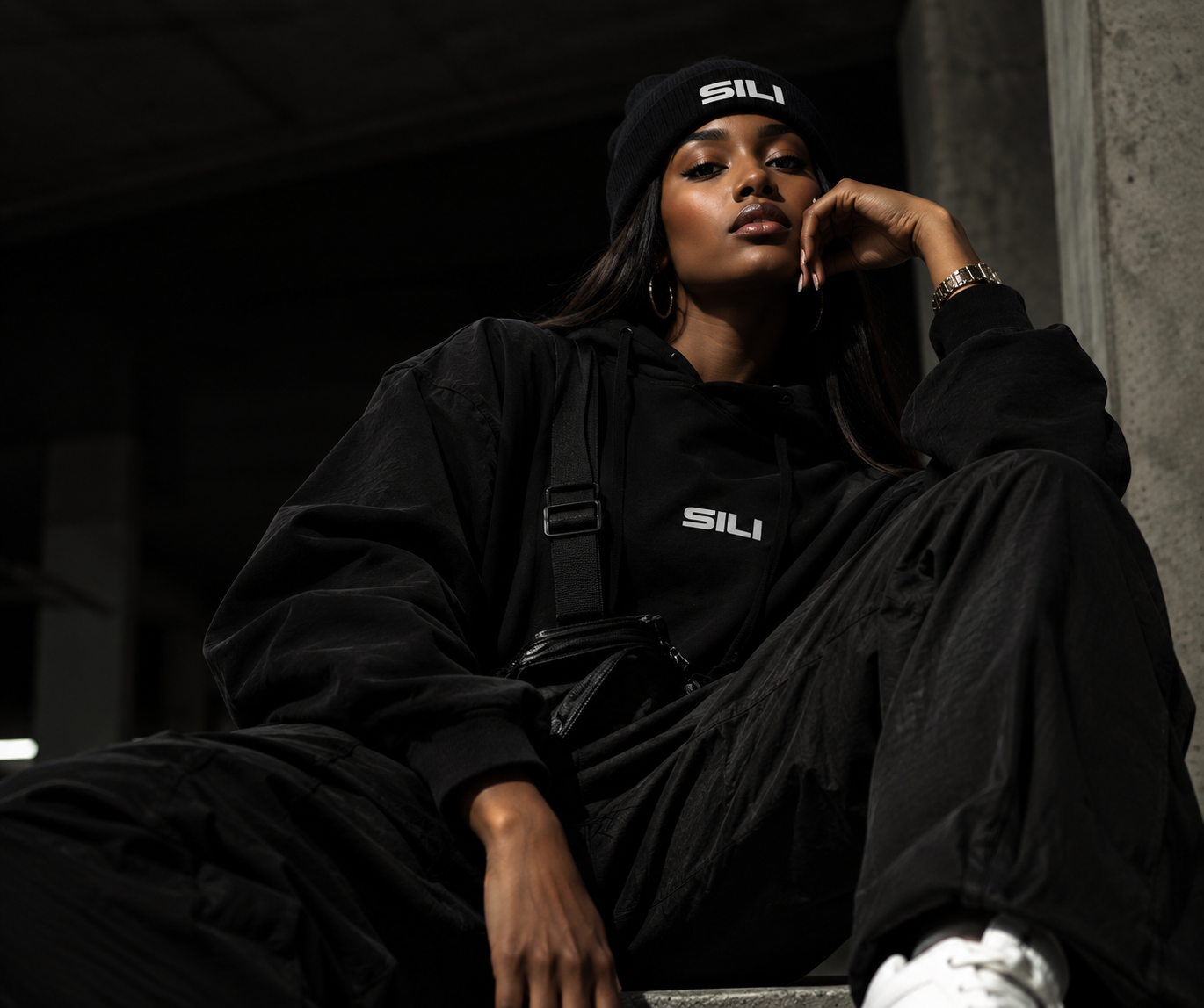 Model wearing an all-black SILI hoodie and beanie against a concrete backdrop